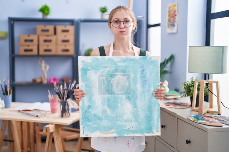 Photo for Young caucasian woman holding canvas at art studio puffing cheeks with funny face. mouth inflated with air, catching air. - Royalty Free Image