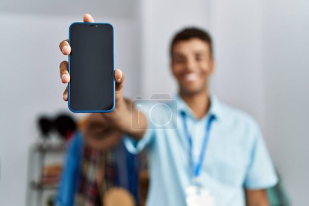 Foto de Joven hispano trabajando como asistente de tienda mostrando la pantalla del teléfono en la tienda minorista - Imagen libre de derechos