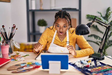 Photo for African american woman with braids sitting at art studio painting looking at tablet scared and amazed with open mouth for surprise, disbelief face - Royalty Free Image
