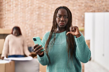 Téléchargez les photos : Femme africaine utilisant smartphone à la nouvelle maison avec le visage en colère, signe négatif montrant aversion avec les pouces vers le bas, concept de rejet - en image libre de droit