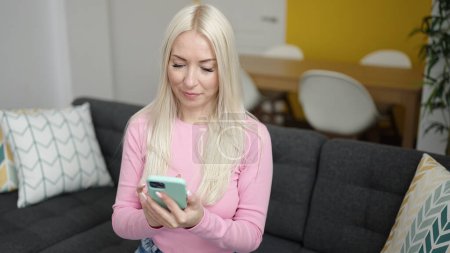 Téléchargez les photos : Jeune femme blonde utilisant un smartphone assis sur un canapé à la maison - en image libre de droit