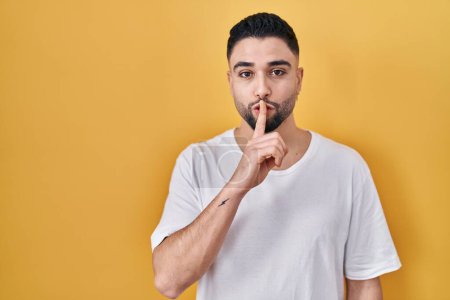 Foto de Young handsome man wearing casual t shirt over yellow background asking to be quiet with finger on lips. silence and secret concept. - Imagen libre de derechos