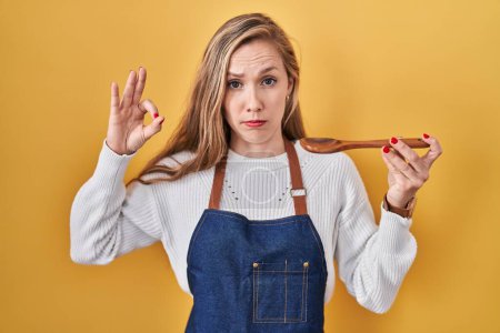 Foto de Young blonde woman wearing apron tasting food holding wooden spoon skeptic and nervous, frowning upset because of problem. negative person. - Imagen libre de derechos