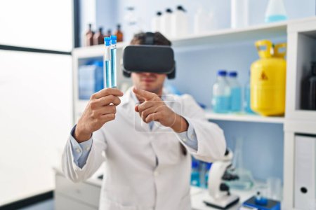 Téléchargez les photos : Jeune homme hispanique scientifique utilisant des lunettes de réalité virtuelle tenant des éprouvettes en laboratoire - en image libre de droit