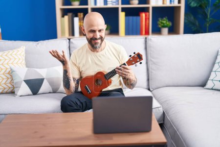 Téléchargez les photos : Jeune homme chauve ayant en ligne ukulele classe assis sur le canapé à la maison - en image libre de droit