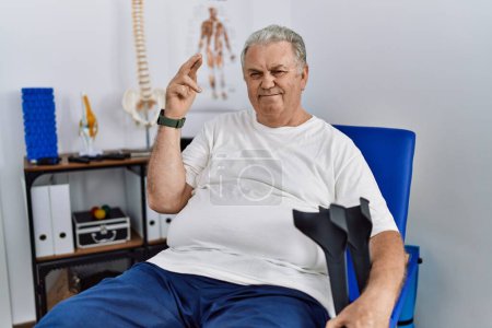 Téléchargez les photos : Senior caucasian man at physiotherapy clinic holding crutches gesturing finger crossed smiling with hope and eyes closed. luck and superstitious concept. - en image libre de droit