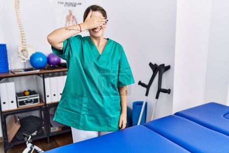 Photo for Young hispanic woman wearing physiotherapist uniform standing at clinic smiling and laughing with hand on face covering eyes for surprise. blind concept. - Royalty Free Image