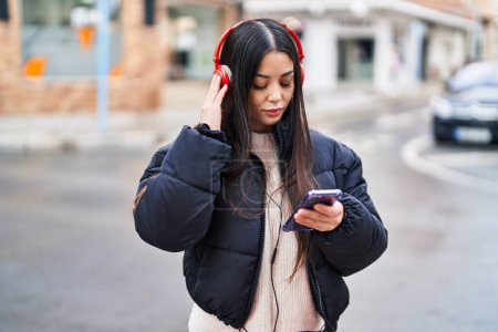 Foto de Joven mujer hispana hermosa escuchando música con expresión relajada en la calle - Imagen libre de derechos