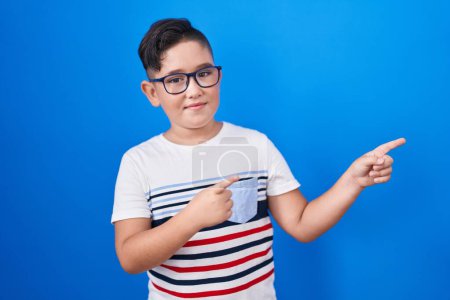 Foto de Joven niño hispano de pie sobre fondo azul sonriendo y mirando a la cámara apuntando con dos manos y dedos hacia un lado. - Imagen libre de derechos