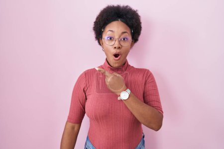 Foto de Beautiful african woman with curly hair standing over pink background surprised pointing with finger to the side, open mouth amazed expression. - Imagen libre de derechos