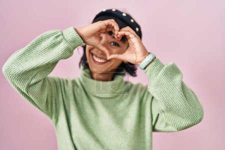 Téléchargez les photos : Jeune belle femme debout sur fond rose faisant forme de coeur avec la main et les doigts souriant en regardant à travers signe - en image libre de droit