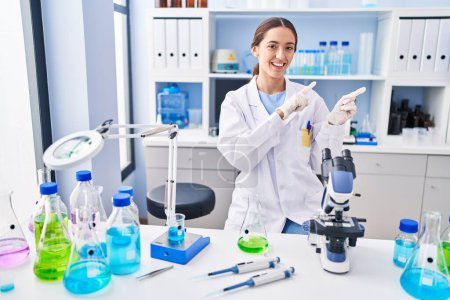 Téléchargez les photos : Young brunette woman working at scientist laboratory smiling and looking at the camera pointing with two hands and fingers to the side. - en image libre de droit