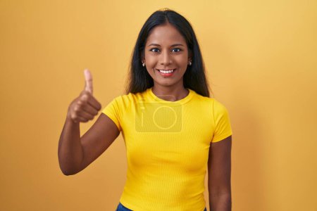 Téléchargez les photos : Jeune femme indienne debout sur fond jaune faisant des pouces heureux geste avec la main. approuvant l'expression regardant la caméra montrant le succès. - en image libre de droit