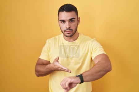 Téléchargez les photos : Jeune homme hispanique debout sur fond jaune à la hâte pointant pour regarder le temps, l'impatience, bouleversé et en colère pour retard de délai - en image libre de droit