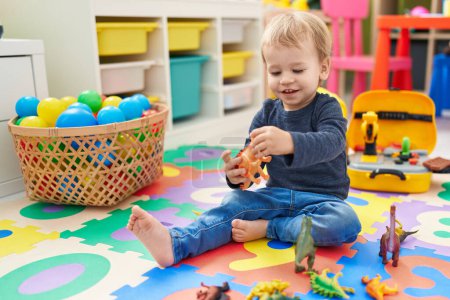Téléchargez les photos : Adorable bambin blond jouant avec des jouets dino assis sur le sol à la maternelle - en image libre de droit