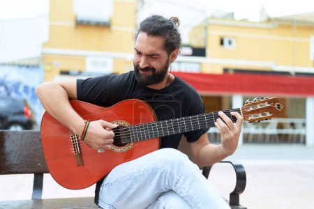 Téléchargez les photos : Jeune homme hispanique musicien jouant de la guitare classique assis sur un banc dans la rue - en image libre de droit