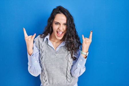 Téléchargez les photos : Jeune femme brune debout sur fond bleu criant avec une expression folle faire symbole de rock avec les mains en l'air. star de la musique. concept de musique lourde. - en image libre de droit