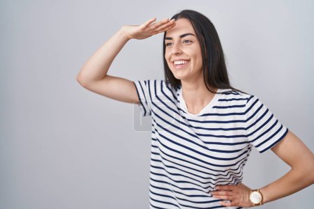 Foto de Young brunette woman wearing striped t shirt very happy and smiling looking far away with hand over head. searching concept. - Imagen libre de derechos