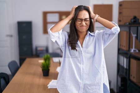 Photo for Young hispanic woman at the office suffering from headache desperate and stressed because pain and migraine. hands on head. - Royalty Free Image