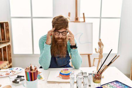 Téléchargez les photos : Redhead man with long beard painting clay bowl at art studio with hand on head for pain in head because stress. suffering migraine. - en image libre de droit