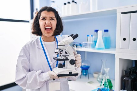 Photo for Young hispanic woman working at scientist laboratory holding microscope angry and mad screaming frustrated and furious, shouting with anger looking up. - Royalty Free Image