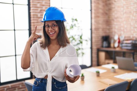 Photo for Hispanic young woman wearing architect hardhat at office smiling happy pointing with hand and finger - Royalty Free Image
