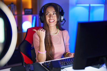 Téléchargez les photos : Young hispanic woman playing video games smiling with happy face looking and pointing to the side with thumb up. - en image libre de droit
