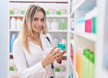 Téléchargez les photos : Jeune femme blonde client souriant confiant tenant bouteille de médicaments à la pharmacie - en image libre de droit