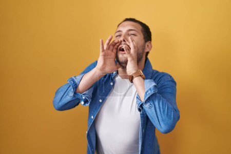 Téléchargez les photos : Hispanic man standing over yellow background shouting angry out loud with hands over mouth - en image libre de droit