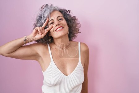 Téléchargez les photos : Middle age woman with grey hair standing over pink background doing peace symbol with fingers over face, smiling cheerful showing victory - en image libre de droit
