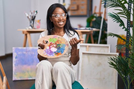 Téléchargez les photos : Jeune femme africaine tenant la palette peintre pointant le pouce vers le côté souriant heureux avec bouche ouverte - en image libre de droit