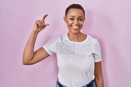 Téléchargez les photos : Beautiful african american woman standing over pink background smiling and confident gesturing with hand doing small size sign with fingers looking and the camera. measure concept. - en image libre de droit