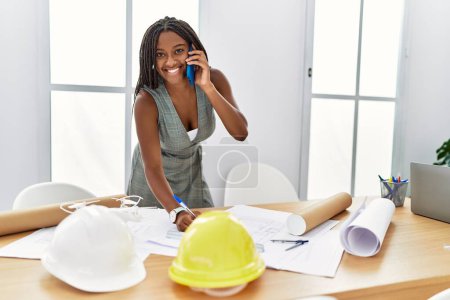 Photo for Young african american woman architect talking on the smartphone writing on house plans at architecture studio - Royalty Free Image