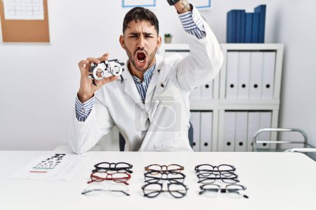 Photo for Young optician man holding optometry glasses angry and mad raising fist frustrated and furious while shouting with anger. rage and aggressive concept. - Royalty Free Image