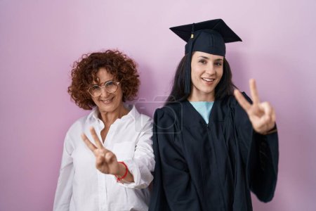 Foto de Madre e hija hispanas con gorra de graduación y bata de ceremonia sonriendo mirando a la cámara mostrando los dedos haciendo señal de victoria. número dos. - Imagen libre de derechos
