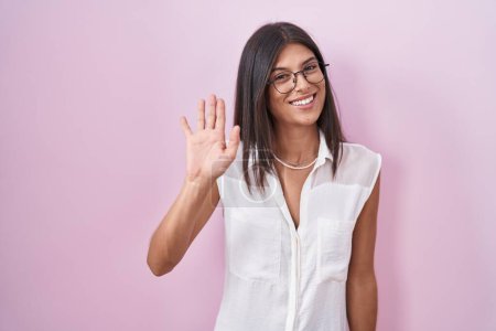 Foto de Brunette young woman standing over pink background wearing glasses waiving saying hello happy and smiling, friendly welcome gesture - Imagen libre de derechos