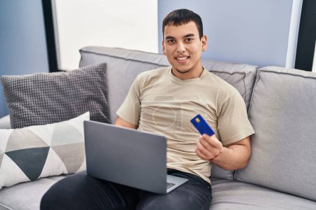 Téléchargez les photos : Jeune homme utilisant un ordinateur portable et carte de crédit assis sur le canapé à la maison - en image libre de droit