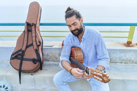 Photo for Young hispanic man musician playing classical guitar sitting on bench at seaside - Royalty Free Image