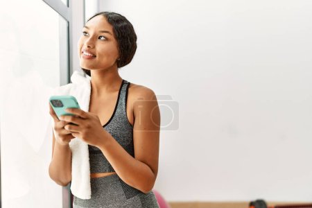 Téléchargez les photos : Jeune femme latine à l'aide d'un smartphone appuyé sur la fenêtre au centre sportif - en image libre de droit