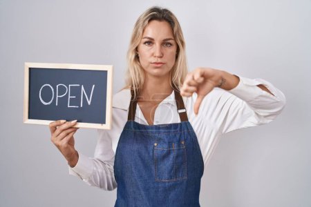 Foto de Young blonde woman wearing apron holding blackboard with open word with angry face, negative sign showing dislike with thumbs down, rejection concept - Imagen libre de derechos