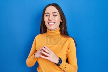 Foto de Young brunette woman standing over blue background hands together and fingers crossed smiling relaxed and cheerful. success and optimistic - Imagen libre de derechos