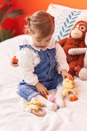 Téléchargez les photos : Adorable bambin blonde jouant avec des jouets assis sur le lit dans la chambre - en image libre de droit