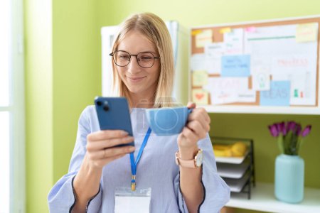 Téléchargez les photos : Jeune femme blonde ouvrière utilisant un smartphone buvant du café au bureau - en image libre de droit