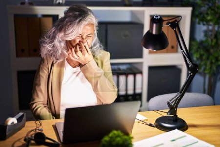 Foto de Middle age woman with grey hair working using computer laptop late at night smelling something stinky and disgusting, intolerable smell, holding breath with fingers on nose. bad smell - Imagen libre de derechos