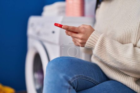 Foto de Young beautiful hispanic woman using smartphone waiting for washing machine at laundry room - Imagen libre de derechos