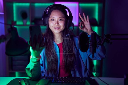 Téléchargez les photos : Young asian woman playing video games with smartphone smiling positive doing ok sign with hand and fingers. successful expression. - en image libre de droit