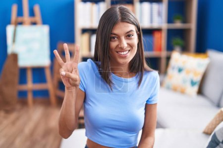 Photo for Brunette young woman sitting on the sofa at home showing and pointing up with fingers number three while smiling confident and happy. - Royalty Free Image