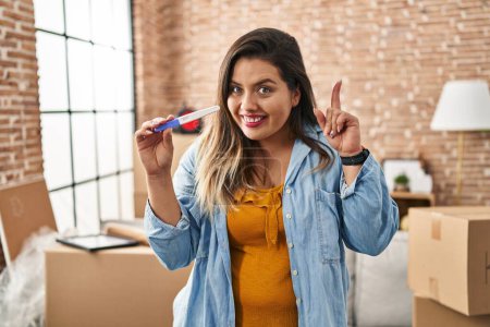Photo for Young hispanic woman holding pregnancy test result at new home surprised with an idea or question pointing finger with happy face, number one - Royalty Free Image