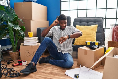 Téléchargez les photos : Afro-Américain assis sur le sol à la nouvelle maison regardant l'heure de la montre inquiet, peur de se faire tard - en image libre de droit