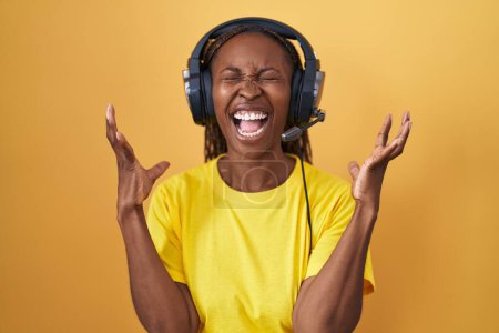 Foto de Mujer afroamericana escuchando música usando auriculares locos y locos gritando y gritando con expresión agresiva y los brazos levantados. concepto de frustración. - Imagen libre de derechos